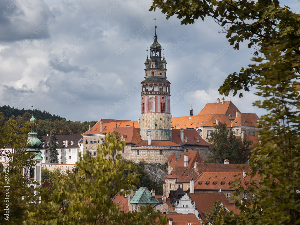 Rooftop view to Cesky Krumlov town. Triangle orange roofs and beautiful bell tower. Cloudy sky, green foliage. No people