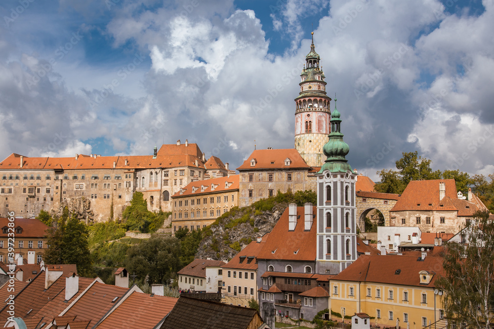 Rooftop view to Cesky Krumlov town. Triangle orange roofs and beautiful bell tower. Cloudy sky, green foliage. No people