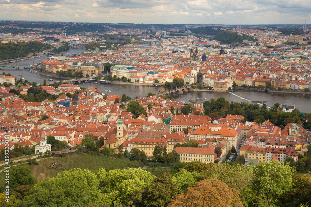 Cityscape of Prague, Czech Republic. View from a hill to the orange tiled roofs and light walls of houses. Green forest in the foreground, river with numerous bridges in the center of the frame.