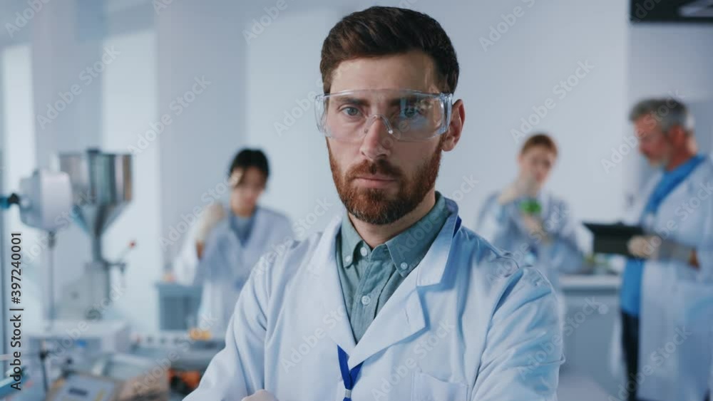 Handsome young man scientist with protective glasses look at camera serious. Background team work. Microbiology pharmaceutical biochemistry medical technology. Slow motion Handsome young man scientist with protective glasses look at camera serious. Background team work. Microbiology pharmaceutical biochemistry medical technology. Slow motion