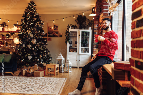 Smiling attractive young bearded man. wearing a kinship sweater, drinking tea from a mug in the living room at home