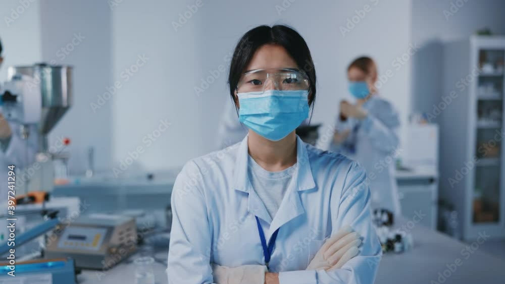 Portrait asian woman scientist with protective glasses and medical mask look at camera. Background team work. Microbiology pharmaceutical biochemistry medical technology. Slow motion Portrait asian woman scientist with protective glasses and medical mask look at camera. Background team work. Microbiology pharmaceutical biochemistry medical technology. Slow motion
