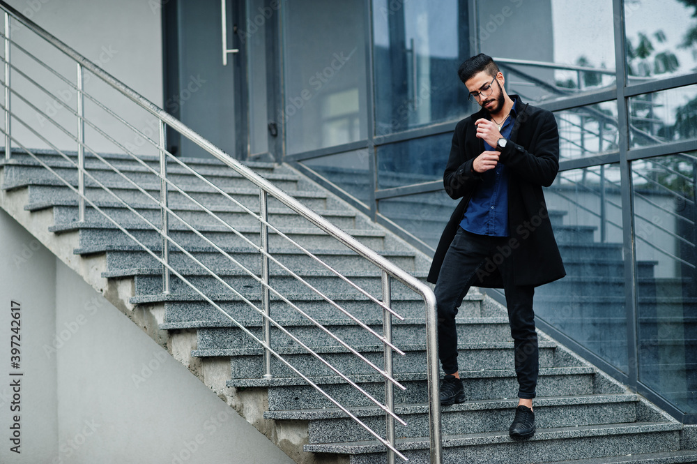 Middle eastern entrepreneur wear black coat and blue shirt, eyeglasses against office building, stand on stairs. Middle eastern entrepreneur wear black coat and blue shirt, eyeglasses against office building, stand on stairs.