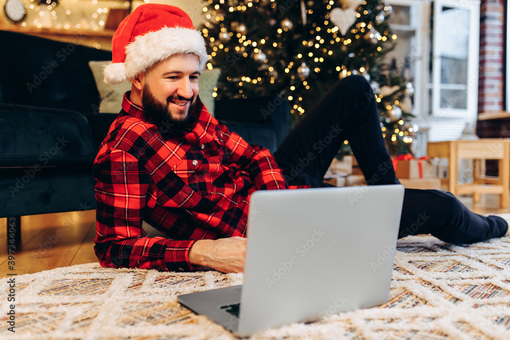 Happy young man with a beard, wearing a Santa Claus hat, uses a laptop, at home against the background of a Christmas tree Happy young man with a beard, wearing a Santa Claus hat, uses a laptop, at home against the background of a Christmas tree