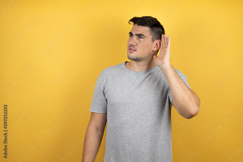 Young handsome man wearing a casual t-shirt over isolated yellow background surprised with hand over ear listening an hearing to rumor or gossip Young handsome man wearing a casual t-shirt over isolated yellow background surprised with hand over ear listening an hearing to rumor or gossip