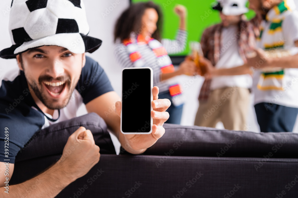 excited football fan showing win gesture while holding smartphone with blank screen near multiethnic friends on blurred background