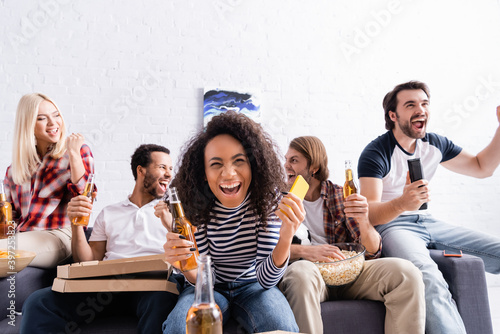 cheerful african american woman holding smartphone and beer while watching football championship with excited multicultural friends