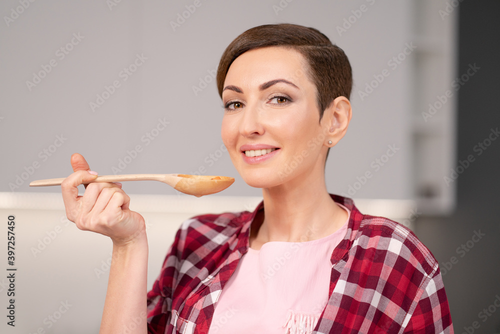Young housewife with a short hairstyle testing a food while prepares dinner for family in the kitchen. Healthy food at home. Healthy food leaving - vegan concept. Young housewife with a short hairstyle testing a food while prepares dinner for family in the kitchen. Healthy food at home. Healthy food leaving - vegan concept.