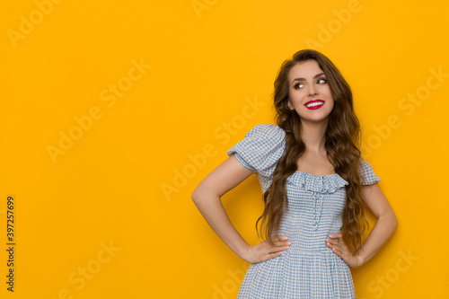 Cheerful Young Woman In Blue Dress Is Posing With Hands On Hip And Looking Away
