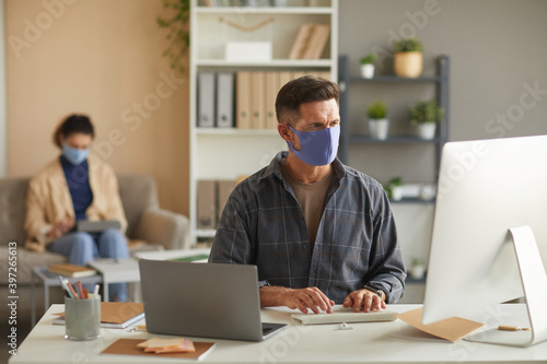 Businessman in protective mask working on computer at his table with his colleague in the background at office