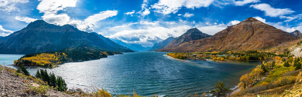 Obraz premium Upper Waterton Lake lakeshore in autumn foliage season sunny day morning. Blue sky, white clouds over mountains in the background. Landmarks in Waterton Lakes National Park, Alberta, Canada.