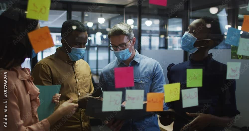 Office colleagues wearing face mask discussing over memo notes on glass board at modern office Office colleagues wearing face mask discussing over memo notes on glass board at modern office