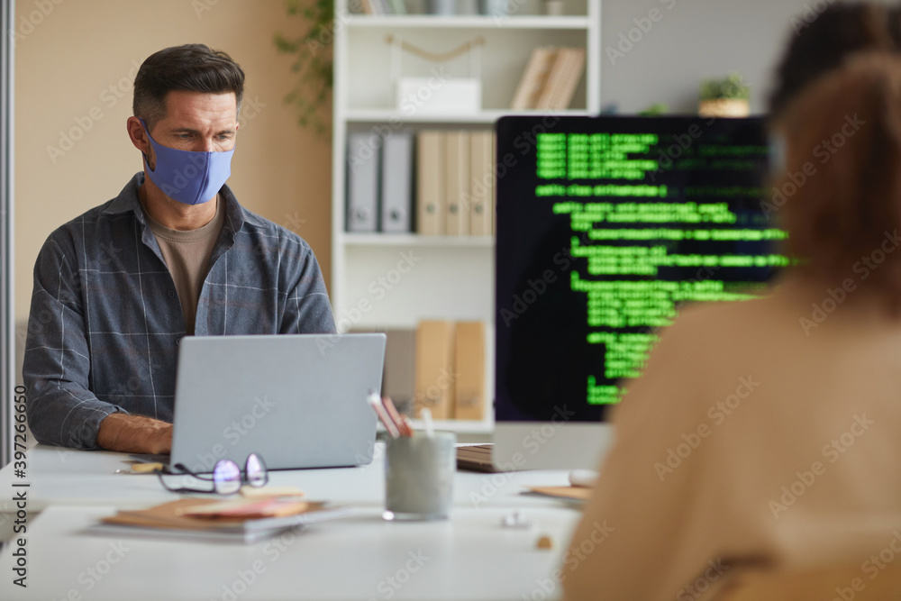 Mature businessman in protective mask working on laptop at the table together with his colleague they working at IT office