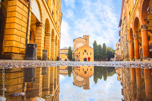 Bologna in the morning, looking Saint Stefano church in square with sunlight and reflection in a puddle on the street. Elderly granmother walking