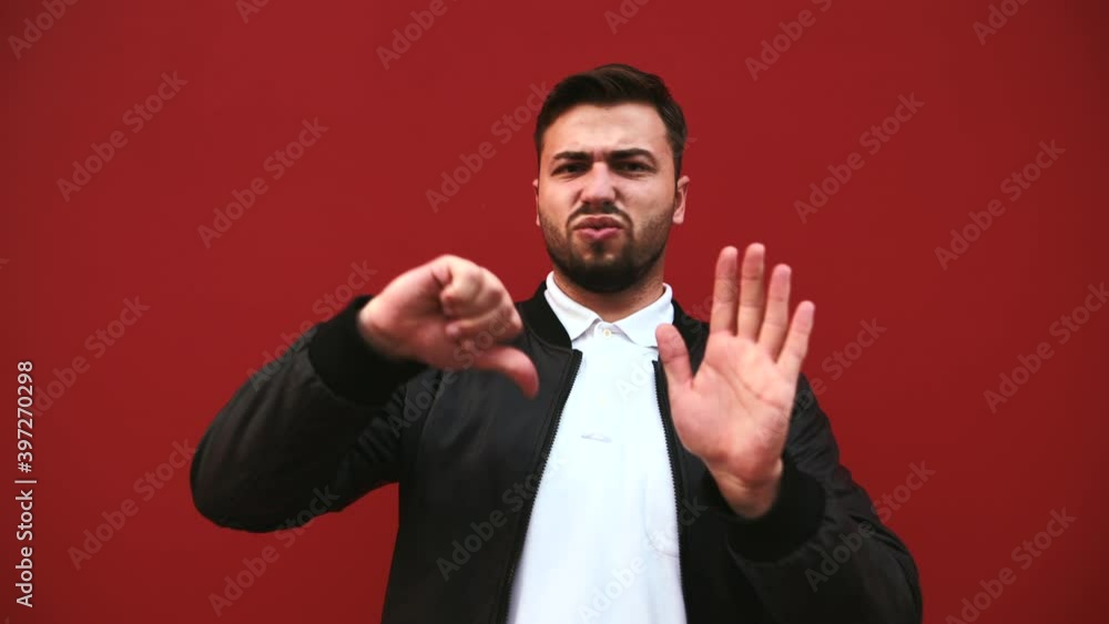 negative bearded man stands against a red wall and shows a dislike gesture with a displeased face looking into the camera. negative bearded man stands against a red wall and shows a dislike gesture with a displeased face looking into the camera.