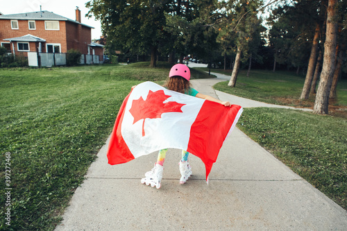 Girl in sport helmet wrapped in large Canadian flag riding on roller skates in park. Canada Day celebration outdoors. Kid in large Canadian flag celebrating national Canada Day on 1 of July.