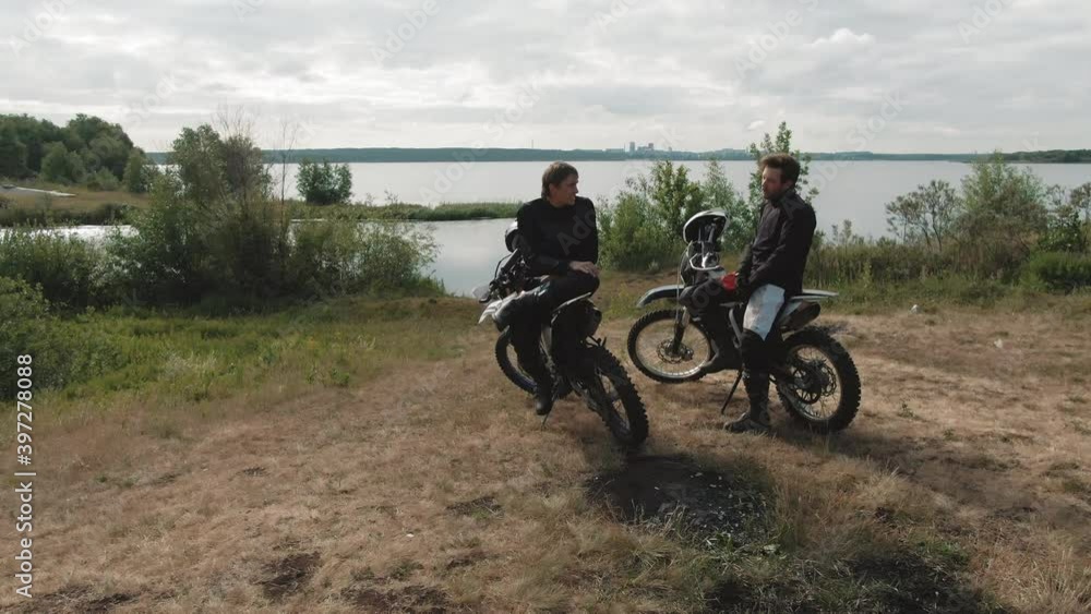 Drone tracking shot of two young men sitting on their motorcycles parked by lake and chatting Drone tracking shot of two young men sitting on their motorcycles parked by lake and chatting