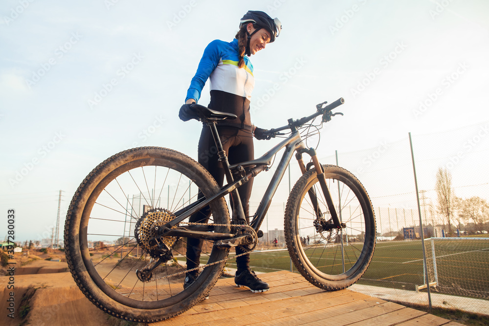 Cheerful young female in sportswear and helmet standing on wooden platform with dusty mountain bike smiling and looking at camera Cheerful young female in sportswear and helmet standing on wooden platform with dusty mountain bike smiling and looking at camera