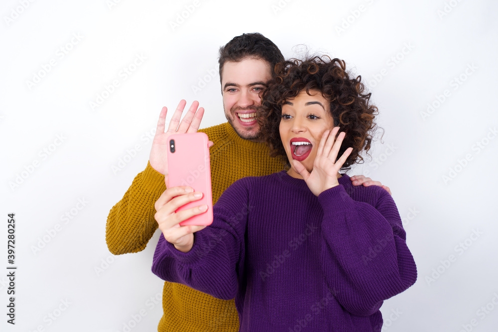 Portrait of happy friendly Young couple wearing knitted sweater standing against white wall taking selfie and waving hand, communicating on video call, online chatting. Portrait of happy friendly Young couple wearing knitted sweater standing against white wall taking selfie and waving hand, communicating on video call, online chatting.