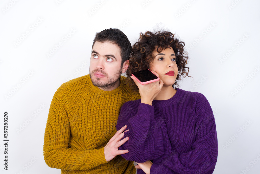 Smiling Young couple wearing knitted sweater standing against white wall listening a voice message from her smartphone. Communication and technology concept. Smiling Young couple wearing knitted sweater standing against white wall listening a voice message from her smartphone. Communication and technology concept.