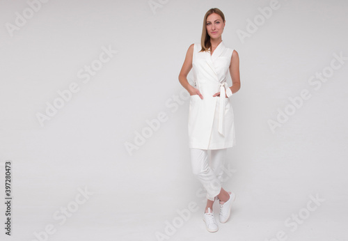 young stylish nurse in white medical costume and white sneakers is standing straight with hands in her pockets and smiling on white wall background. medical concept. free space on left side