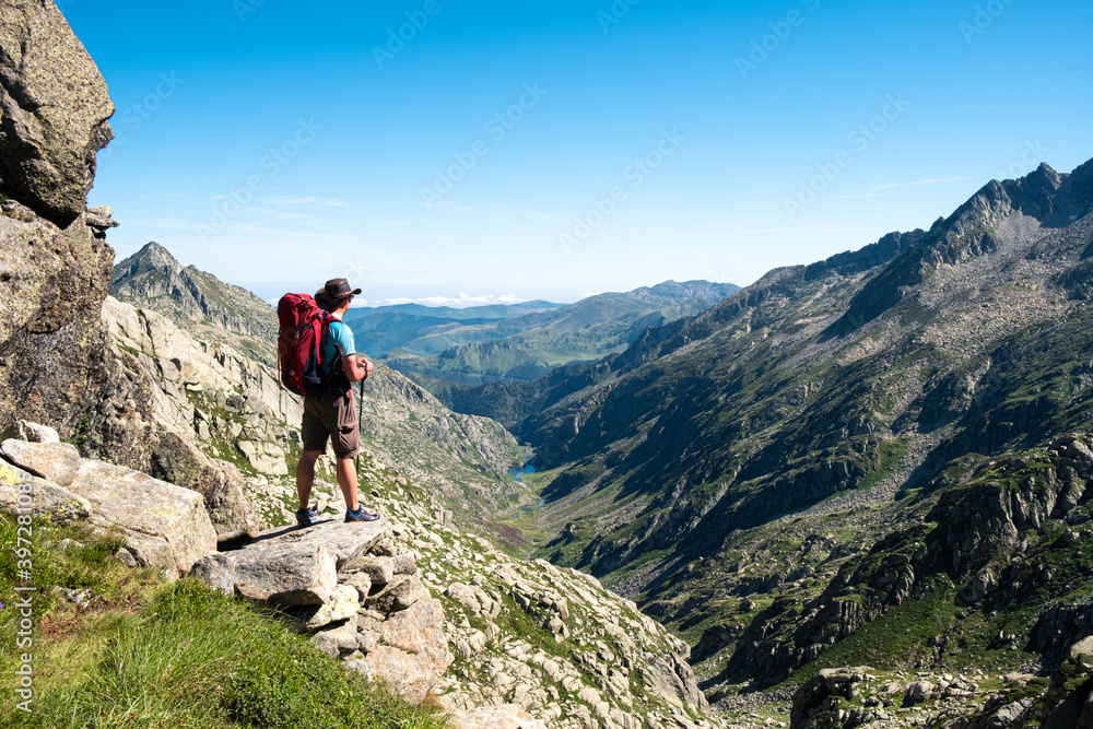 Back view of unrecognizable male hiker with backpack and trekking stick standing on rocky hilltop and admiring spectacular scenery of mountain ridge in summer day Back view of unrecognizable male hiker with backpack and trekking stick standing on rocky hilltop and admiring spectacular scenery of mountain ridge in summer day