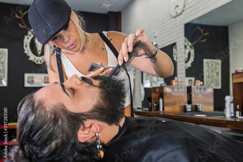 Side view of stylish tattooed female master shaving bearded customer with electric machine while working in professional barbershop photo