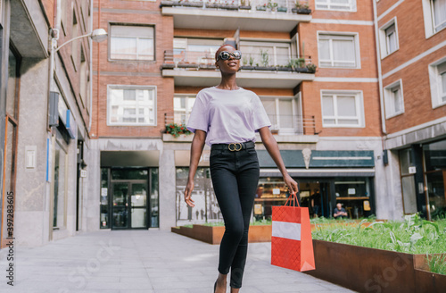 Wallpaper Mural Full body of confident stylish young African American female shopaholic in trendy clothes and sunglasses with shopping bag in hand walking on street near fashion store Torontodigital.ca