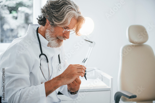 Portrait of senior male doctor with grey hair and beard in white coat is indoors in otorhinolaryngology clinic
