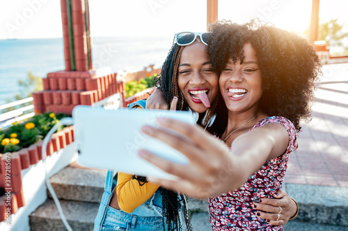 Smiling black woman with braids hugging cheerful African American female friend with curly hair and taking selfie together while standing on promenade in summer