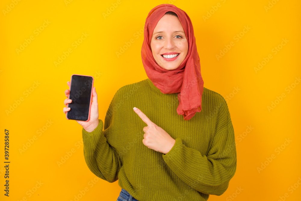 Smiling Young Caucasian Muslim woman wearing hijab standing against yellow wall showing and pointing at empty phone screen. Advertisement and communication concept. Smiling Young Caucasian Muslim woman wearing hijab standing against yellow wall showing and pointing at empty phone screen. Advertisement and communication concept.