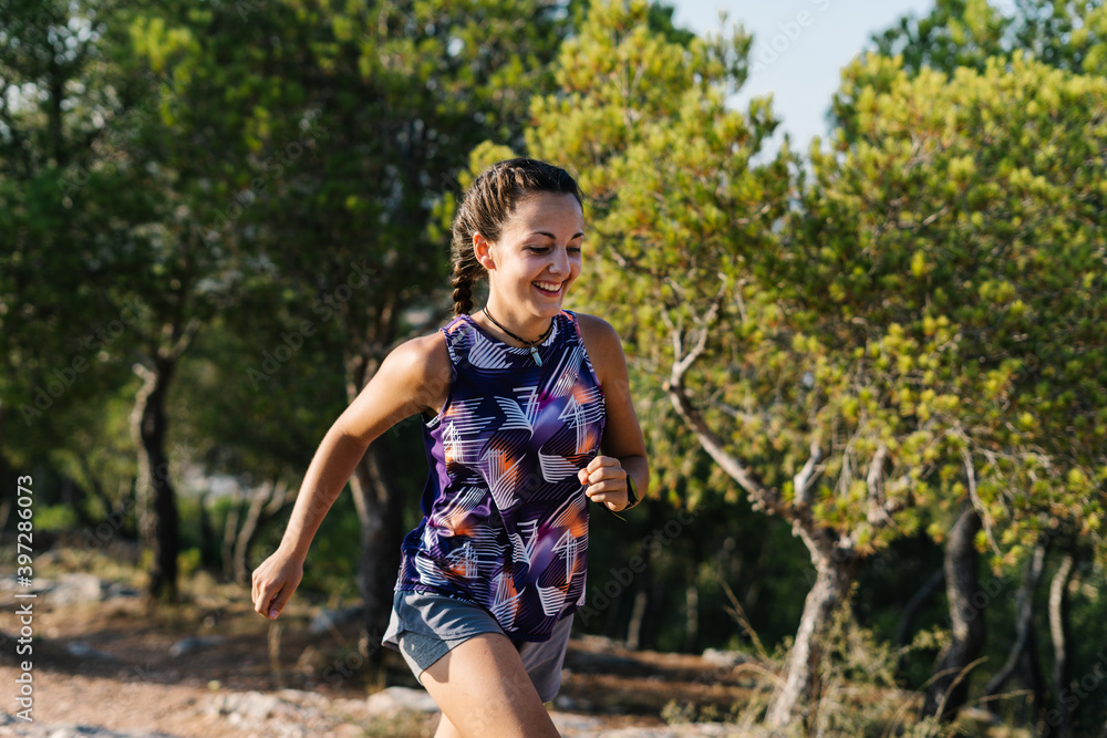 Positive young athletic female in activewear running on stony trail in green forested mountainous terrain Positive young athletic female in activewear running on stony trail in green forested mountainous terrain