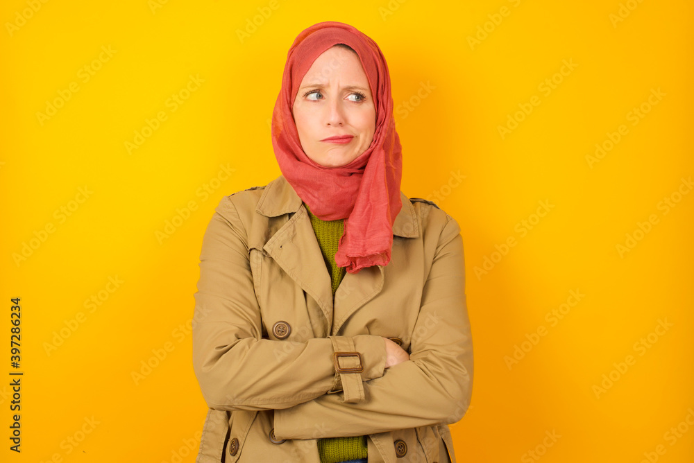 Picture of angry Young Caucasian Muslim woman wearing hijab standing against yellow wall crossing arms. Looking at camera with disappointed expression. Picture of angry Young Caucasian Muslim woman wearing hijab standing against yellow wall crossing arms. Looking at camera with disappointed expression.