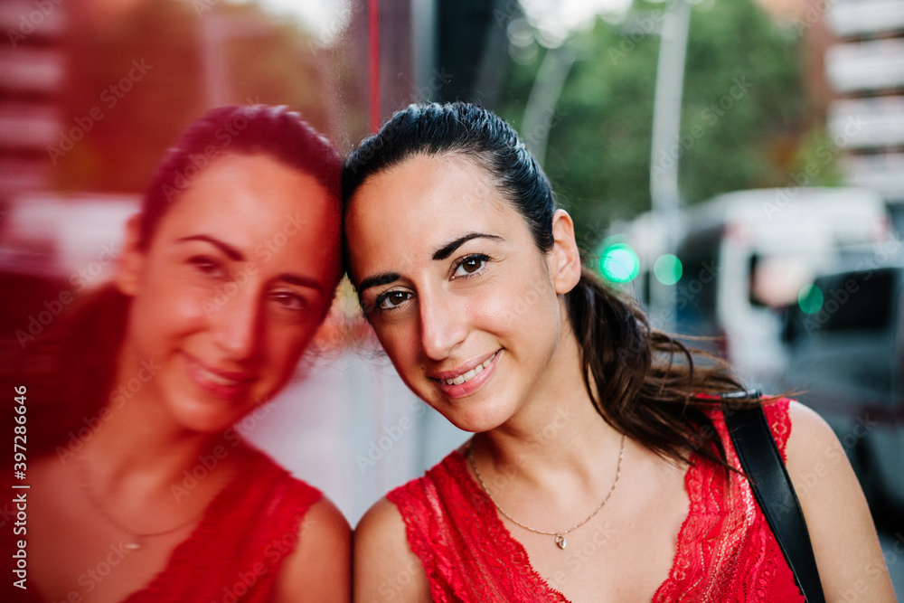 Cheerful young Hispanic female in red blouse leaning on glass wall of modern building and looking at camera while standing on urban street Cheerful young Hispanic female in red blouse leaning on glass wall of modern building and looking at camera while standing on urban street