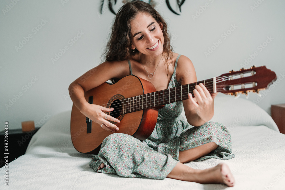 Cheerful ethnic female guitarist sitting on cozy bed at home and playing acoustic musical instrument with closed eyes Cheerful ethnic female guitarist sitting on cozy bed at home and playing acoustic musical instrument with closed eyes