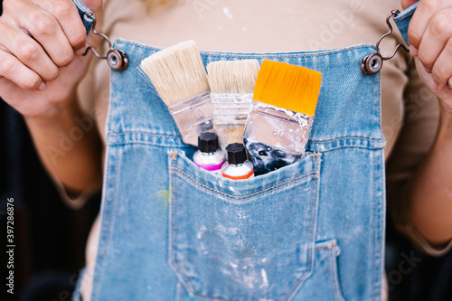 Crop anonymous female artist in denim overall with various paintbrushes and colorful paint tubes in pocket photo