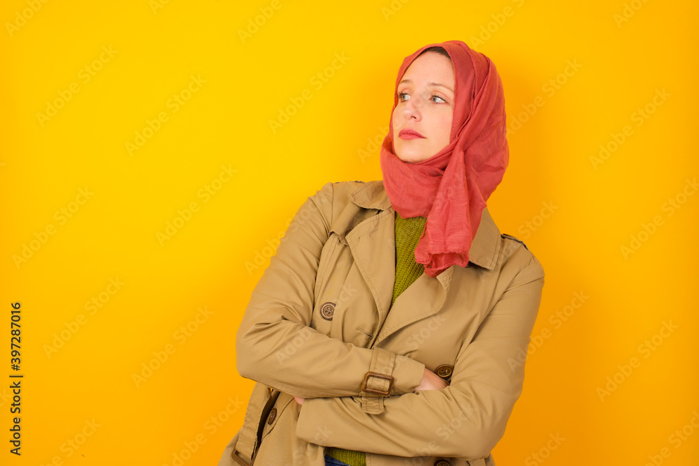 Image of upset Young caucasian Muslim woman wearing hijab standing against yellow wall with arms crossed. Looking with disappointed expression aside after listening to bad news. Image of upset Young caucasian Muslim woman wearing hijab standing against yellow wall with arms crossed. Looking with disappointed expression aside after listening to bad news.