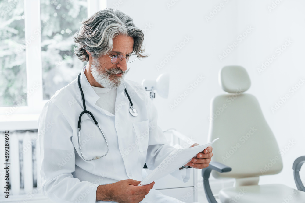 Portrait of senior male doctor with grey hair and beard in white coat is indoors in clinic Portrait of senior male doctor with grey hair and beard in white coat is indoors in clinic