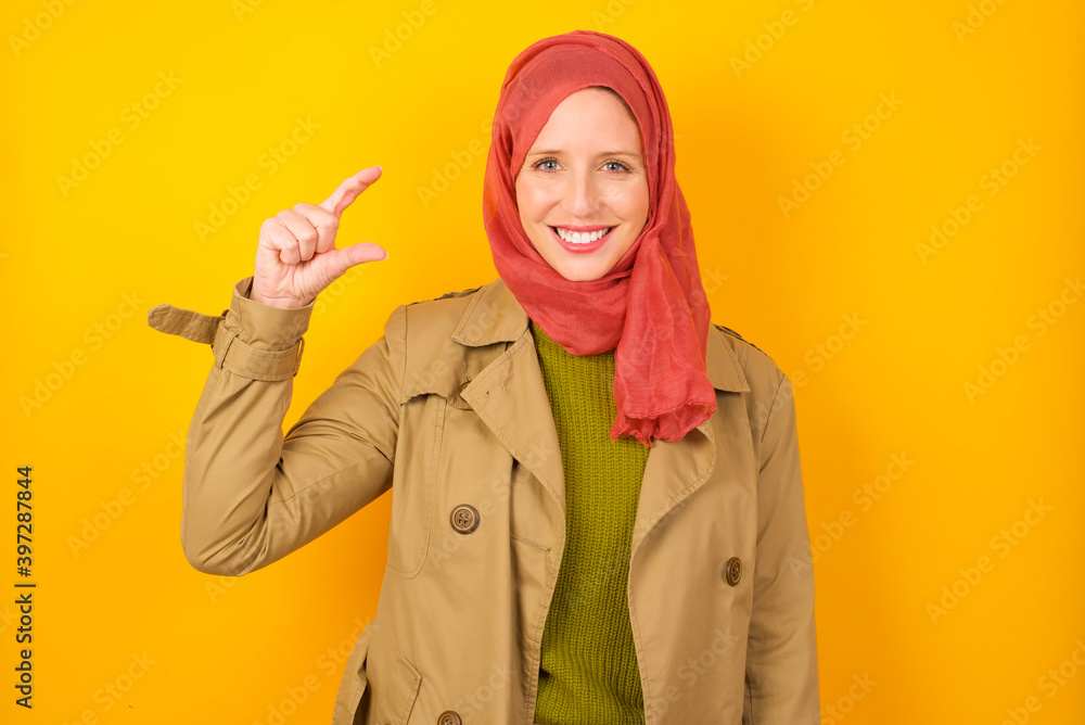 Young caucasian Muslim woman wearing hijab standing against yellow wall smiling and gesturing with hand small size, measure symbol. Young caucasian Muslim woman wearing hijab standing against yellow wall smiling and gesturing with hand small size, measure symbol.