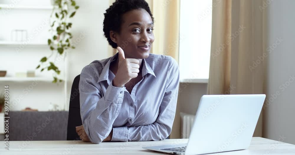 Afro american student black business woman sitting at workplace home office looking at laptop computer screen reads good news shows on camera like puts thumbs up makes gesture of success approval Afro american student black business woman sitting at workplace home office looking at laptop computer screen reads good news shows on camera like puts thumbs up makes gesture of success approval