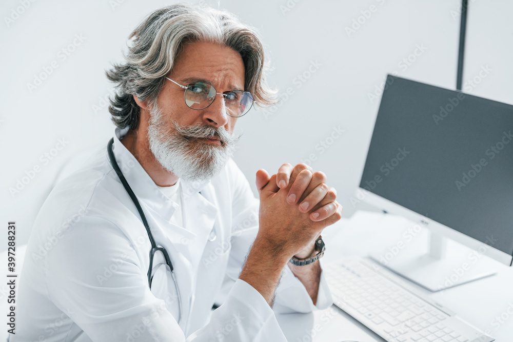 Portrait of senior male doctor with grey hair and beard in white coat is indoors in clinic Portrait of senior male doctor with grey hair and beard in white coat is indoors in clinic