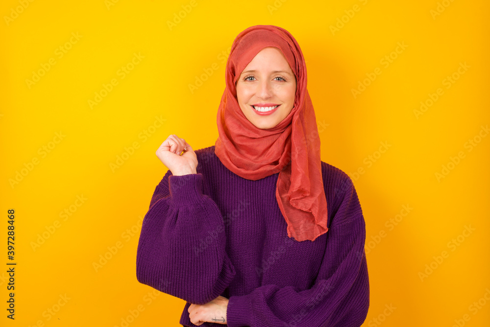 Cheerful Young caucasian Muslim woman wearing hijab standing against yellow wall with hand near face. Looking with glad expression at the camera after listening to good news. Confidence. Cheerful Young caucasian Muslim woman wearing hijab standing against yellow wall with hand near face. Looking with glad expression at the camera after listening to good news. Confidence.