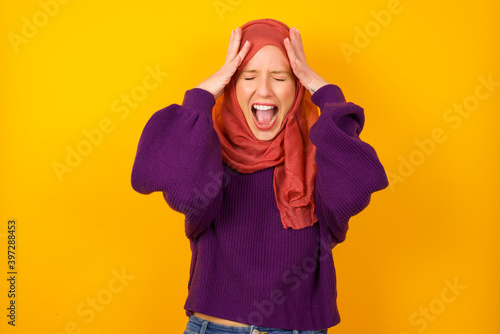 Shocked panic Young caucasian Muslim woman wearing hijab standing against yellow wall holding hands on head and screaming in despair and frustration. © Jihan