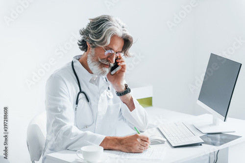 Writes by computer table. Senior male doctor with grey hair and beard in white coat is indoors in clinic