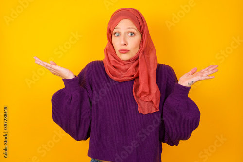 Puzzled and clueless Young caucasian Muslim woman wearing hijab standing against yellow wall with arms out, shrugging shoulders, saying: who cares, so what, I don't know.