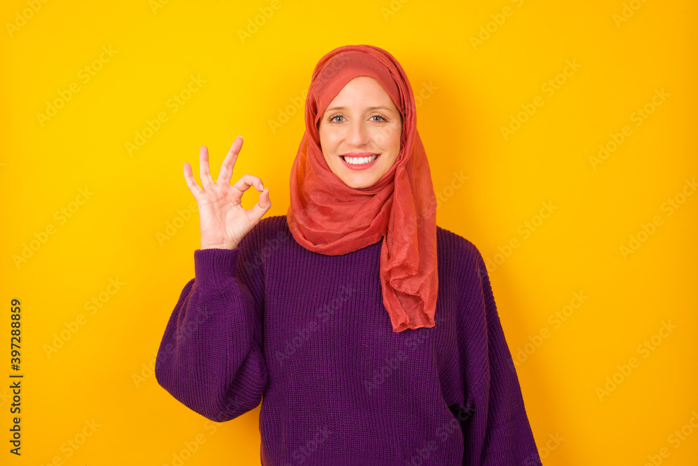 Glad attractive Young caucasian Muslim woman wearing hijab standing against yellow wall shows ok sign with hand as expresses approval, has cheerful expression, being optimistic. Glad attractive Young caucasian Muslim woman wearing hijab standing against yellow wall shows ok sign with hand as expresses approval, has cheerful expression, being optimistic.