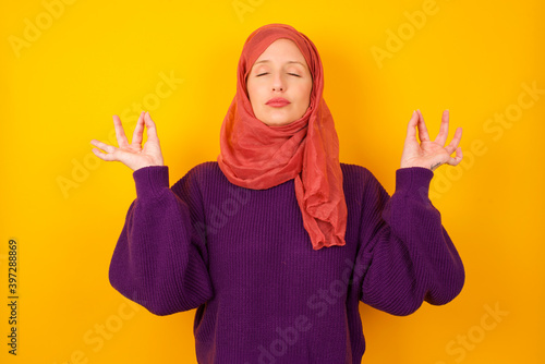 Young caucasian Muslim woman wearing hijab standing against yellow wall doing yoga, keeping eyes closed, holding fingers in mudra gesture. Meditation, religion and spiritual practices. photo