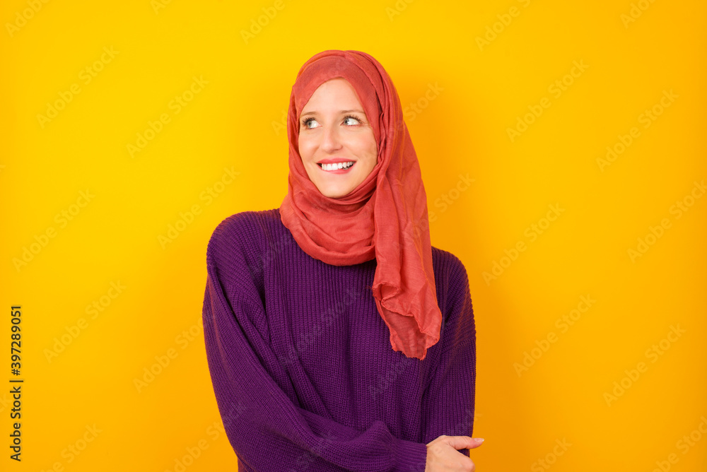 Young caucasian Muslim woman wearing hijab standing against yellow wall with thoughtful expression, looks away keeps hands down bitting his lip thinks about something pleasant. Young caucasian Muslim woman wearing hijab standing against yellow wall with thoughtful expression, looks away keeps hands down bitting his lip thinks about something pleasant.