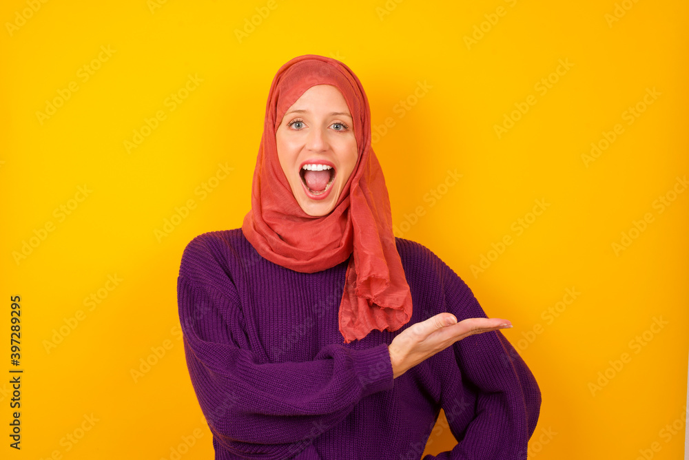 Young caucasian Muslim woman wearing hijab standing against yellow wall says: wow how exciting it is, has amazed expression, shows something on blank space with palm. Advertisement concept. Young caucasian Muslim woman wearing hijab standing against yellow wall says: wow how exciting it is, has amazed expression, shows something on blank space with palm. Advertisement concept.