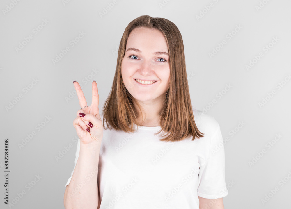Young smiling woman stands in a white T-shirt on a gray background and shows a peace sign. Young smiling woman stands in a white T-shirt on a gray background and shows a peace sign.